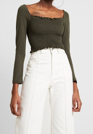 Vrouw die een olijfgroen cropped top met lange mouwen en plooien draagt, gecombineerd met een witte broek met hoge taille, met een rozentattoo zichtbaar op de bovenste borst.