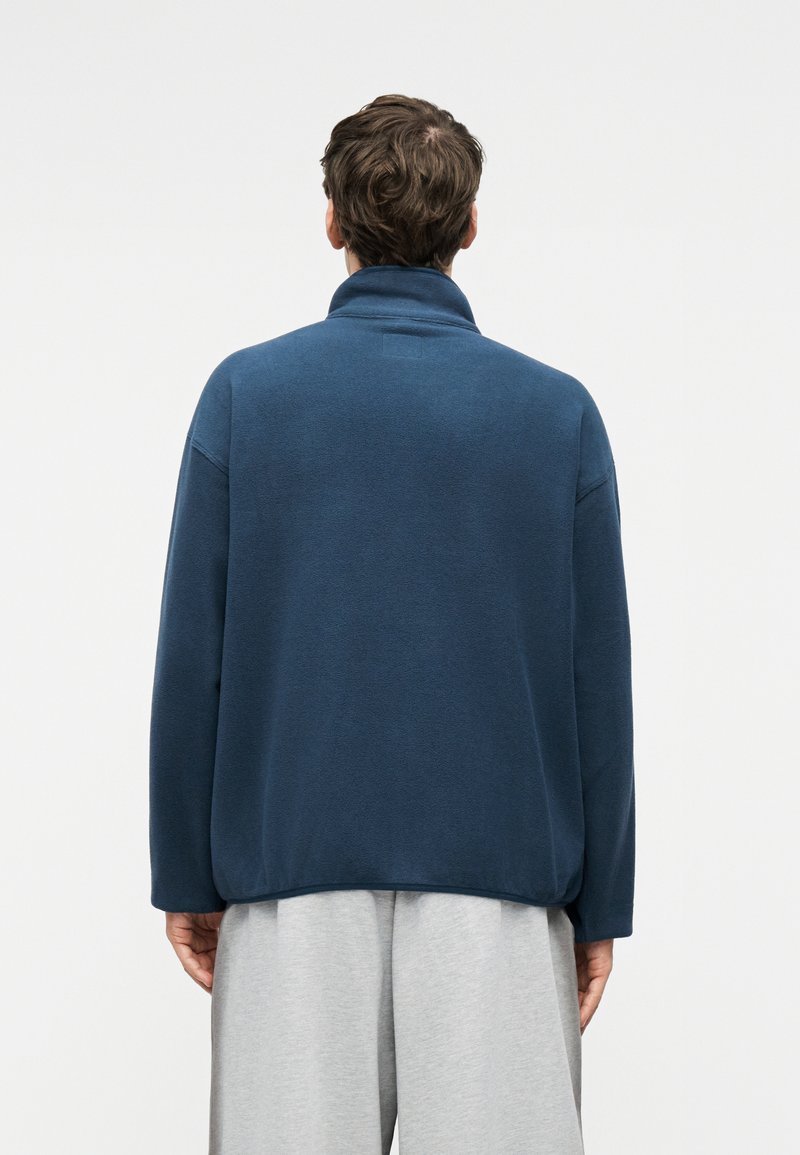 Man with short brown hair wearing a navy blue fleece jacket and light gray pants, standing facing away against a plain background.