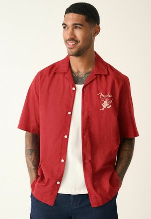 Man with tattoos wearing a red short-sleeve button-up shirt over a white tank top, smiling and looking to the side.