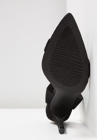 Chaussure noire à talon haut et pointe pointue reposant contre un mur d’angle blanc, montrant une semelle texturée et un talon fin.