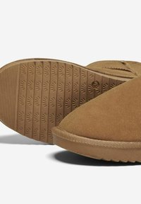 Zapatos de ante marrón tipo slip-on con suela de goma texturizada que presenta un agarre con patrón. Los acentos cosidos a lo largo de los bordes completan el diseño.