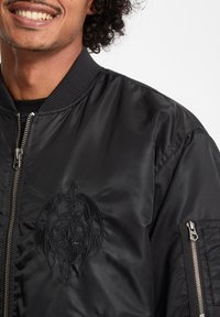 Giacca bomber nera con colletto a coste, tasche con zip e un design grafico ricamato sul lato sinistro. Materiale liscio e lucido.