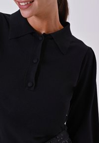 Top nero a maniche lunghe con colletto, dotato di tre bottoni frontali e tessuto morbido, abbinato a una cintura nera con paillettes.