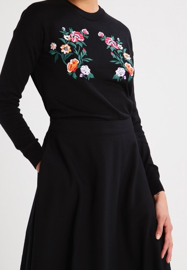 Musta pitkähihainen paita, jossa on punaisen, oranssin, vaaleanpunaisen ja vihreän kukka-aiheinen kirjailu, yhdistettynä mustaan, täyspitkään ja keskipitkään hameeseen.