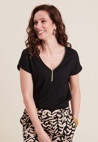 Top nero a maniche corte con scollatura a V e rifiniture dorate. Presenta un tessuto texturizzato ed è abbinato a pantaloni beige e neri con motivo.