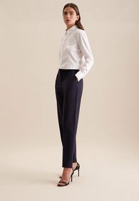 Camicia bianca ritagliata con colletto, abbinata a pantaloni blu navy a vita alta e sandali con tacco a spillo neri. Tessuto liscio, vestibilità su misura, dettagli minimalisti.