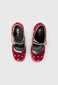 Παπούτσια Mary Jane από κόκκινο βελούδο με χρυσό λουράκι και διακοσμητικά πετράδια, με λείο μαύρο εσωτερικό πέλμα και διακοσμητικά καρφιά στο επάνω μέρος.