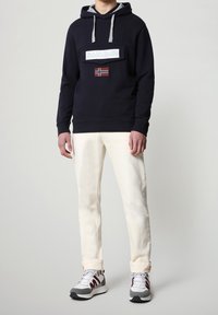 Felpa con cappuccio navy con accenti grigi, dotata di un'etichetta con logo bianco e un patch della bandiera della Norvegia. Abbinata a pantaloni crema e sneakers grigie.