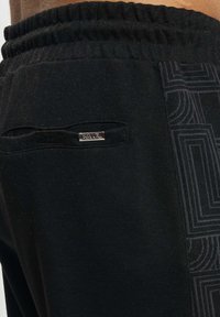 Svarte joggere laget av mykt stoff har en ribbet midje, logomerke og et teksturert panel med et geometrisk mønster på siden.
