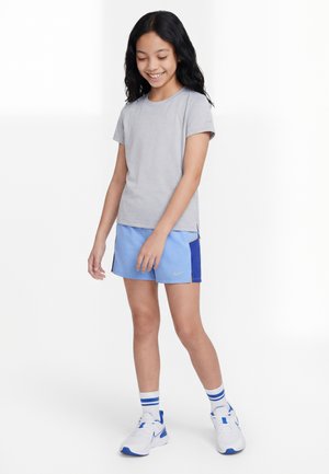 Djevojka se smiješi i gleda dolje, nosi sivu majicu, svijetloplave sportske kratke hlače, bijele tenisice i bijele čarape s plavim prugama.