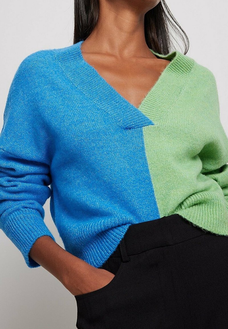 Suéter con diseño de bloques de color y cuello en V, con una mitad en azul brillante y la otra en verde claro; tejido suave y texturizado.