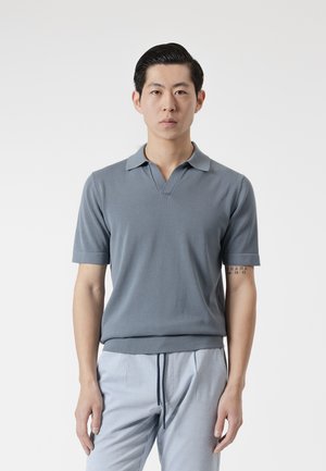 Hombre con cabello corto y negro, vistiendo una camiseta polo gris de manga corta y pantalones azules claros con cordón, de pie frente a un fondo liso.