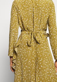 Bakvy av person som bär en senapsgul klänning med vitt blommönster, långa ärmar och en knuten tygbälte i midjan.