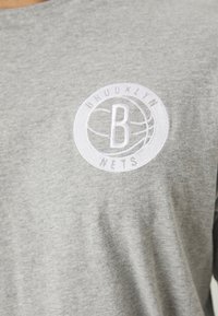 Camiseta de algodón gris con un logo circular bordado en blanco que dice "BROOKLYN" en la parte superior y "NETS" en la parte inferior, centrado en el pecho.