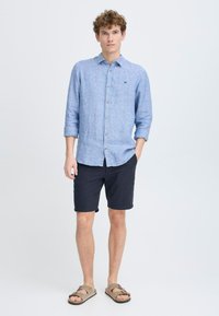 Lys blå linne skjorte med knapper og lange ermer, kombinert med mørke shorts og brune sandaler. Skjorten har en subtil tekstur og krage.