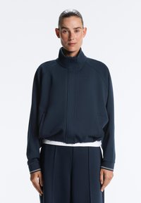 Kvinde står med fronten mod kamera iført en marineblå lynlåsjakke med høj krave og matchende marineblå bukser mod en ensfarvet hvid baggrund.