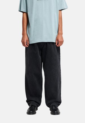 Osoba nosząca luźne czarne jeansy, czarne buty oraz jasnoniebieski oversize T-shirt, stojąca na jednolitym tle.