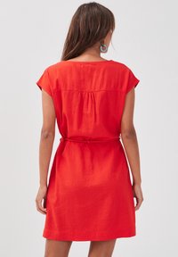 Robe en lin rouge avec de courtes manches à capuche, des détails froncés au dos et une taille nouée. Texture lisse, longueur au genou.