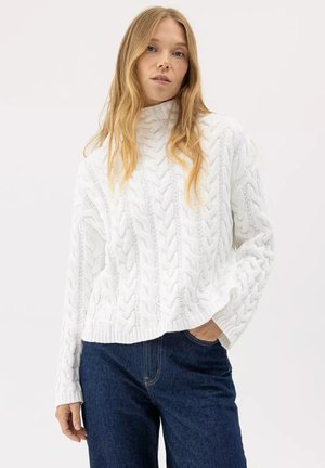 Donna bionda che indossa un maglione bianco a collo alto lavorato a trecce e jeans blu scuro, in piedi con una mano in tasca.