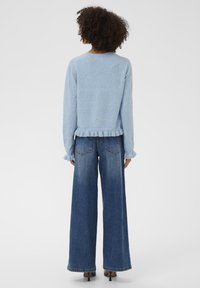 Göndör hajú nő, világoskék, fodros szegélyű pulóvert, bő szárú kék farmert és fekete magassarkú cipőt visel, háttal állva.