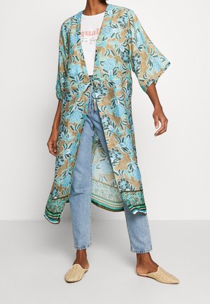 Personne portant un kimono fleuri bleu et beige sur un t-shirt blanc graphique, un jean bleu clair et des chaussures beige tissées à enfiler.