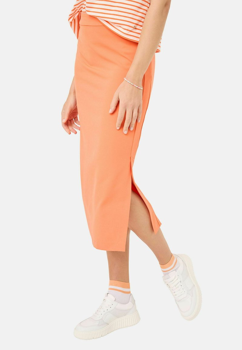 Jupe midi orange avec des fentes latérales, texture lisse et design ajusté, associée à des sneakers blanches et des chaussettes rayées.