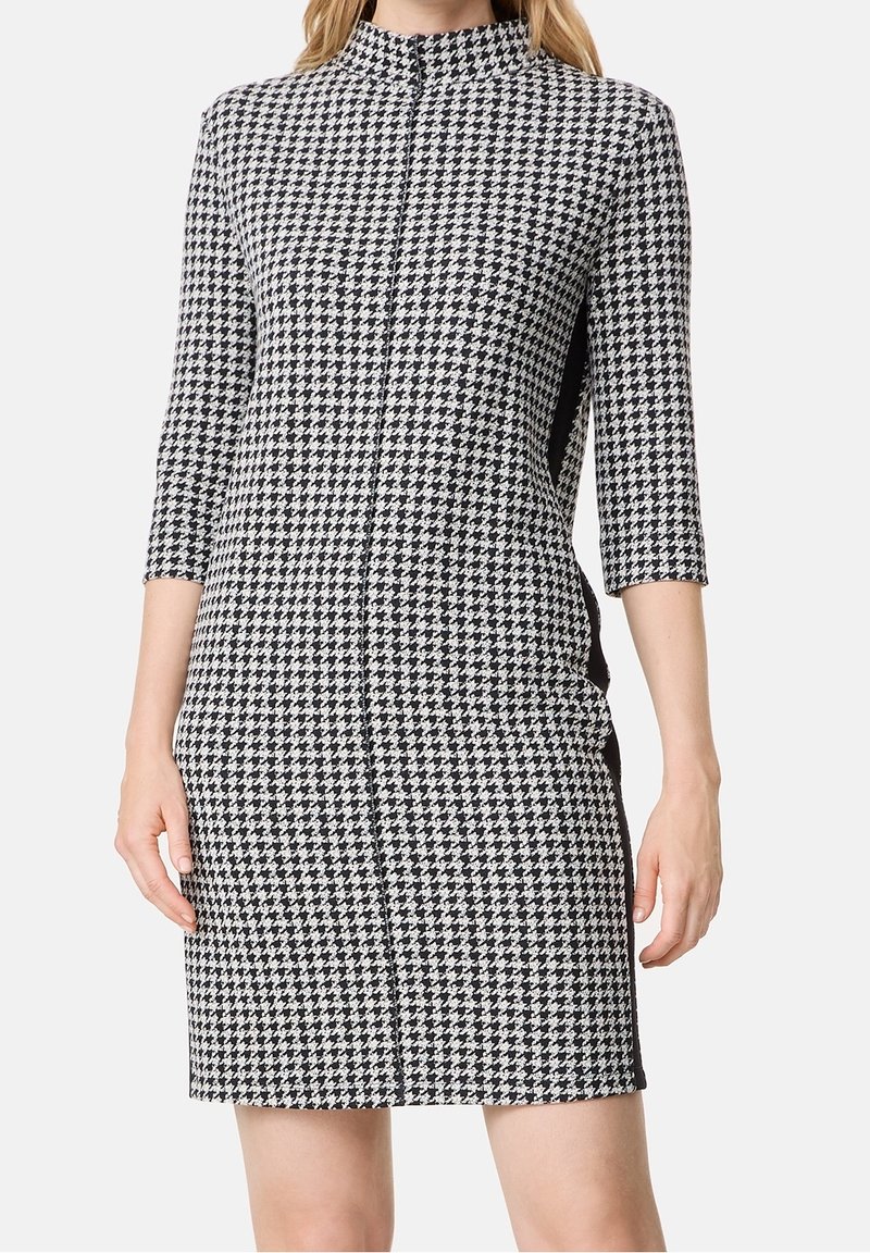 Femme portant une robe à motif pied-de-poule noir et blanc avec des manches trois-quarts et un col montant, debout devant un fond blanc.