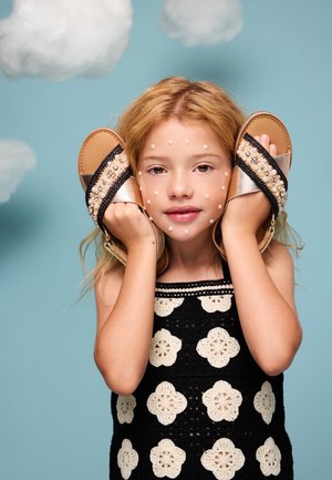 Joven con decoraciones de perlas en el rostro sostiene sandalias adornadas contra sus orejas, sobre un fondo azul con nubes esponjosas.