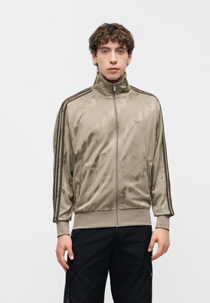 Giovane uomo con capelli corti e ricci che indossa una giacca beige con zip e strisce scure sulle maniche e pantaloni neri, in piedi davanti a uno sfondo chiaro uniforme.