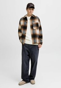 Brown and black plaid oversized flannel shirt, white knit sweater, dark jeans, and beige moccasin-style shoes. Cap with “Originals” text.