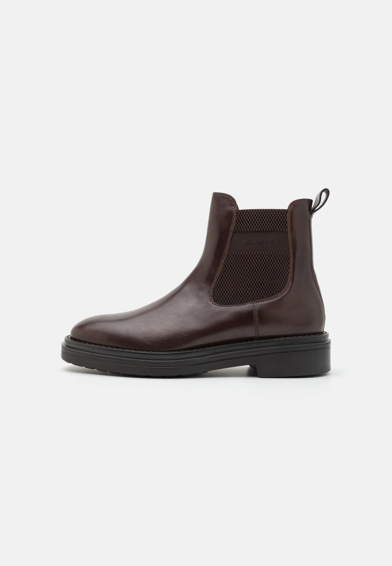 Botas Chelsea de cuero marrón con paneles laterales elásticos, puntera redonda y suela de goma negra gruesa. Textura suave y detalles mínimos.