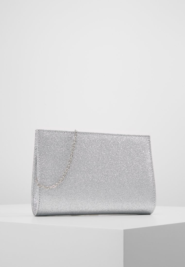 Pochette à paillettes argentées avec une fine chaîne, posée sur un socle blanc sur un fond gris clair uni.