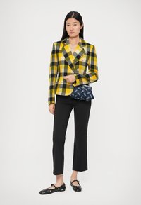 Κίτρινο και μαύρο καρό σακάκι με διπλά κουμπιά, συνδυασμένο με μια μοτίβο ναυτική τσάντα, μαύρο παντελόνι και μαύρες φλατ παπούτσια.