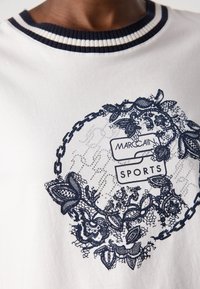 T-shirt in cotone bianco con un design in pizzo floreale blu navy, scollatura a costine con righe blu e bianche, e logo "Marc Cain Sports".
