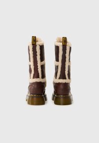 Brune læder ankelstøvler med shearling-fôr, gule sting og en karakteristisk gul træksløjfe bagpå. Rund toe og tyk sål.