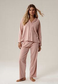 Kobieta z długimi blond włosami stoi boso, ubrana w luźny różowy piżama, składający się z koszulki zapinanej na guziki i spodni z małymi białymi wzorami w kształcie półksiężyca.