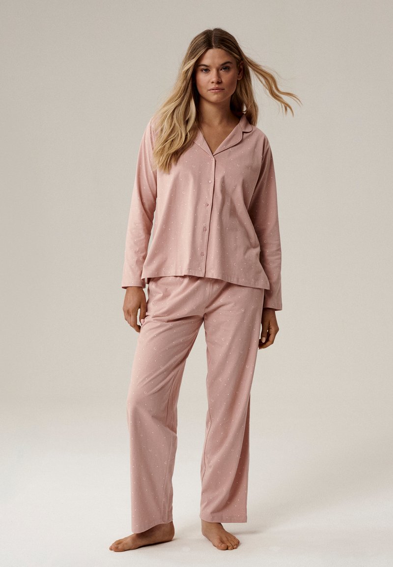 Kobieta z długimi blond włosami stoi boso, ubrana w luźny różowy piżama, składający się z koszulki zapinanej na guziki i spodni z małymi białymi wzorami w kształcie półksiężyca.