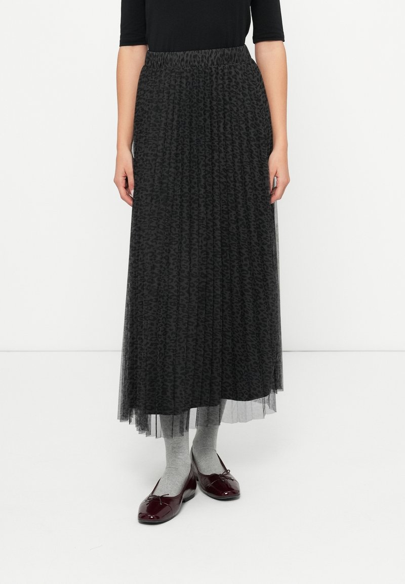 Femme portant une longue jupe noire plissée à imprimé animal, des chaussettes grises, des ballerines vernis bordeaux et un haut noir, debout sur un sol blanc.