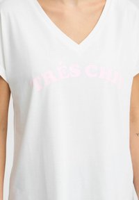 Balta kokvilnas t-krekls ar v veida kaklu un īsām piedurknēm, rotāts ar rozā tekstu "TRÈS CHIC" krūšu daļā.