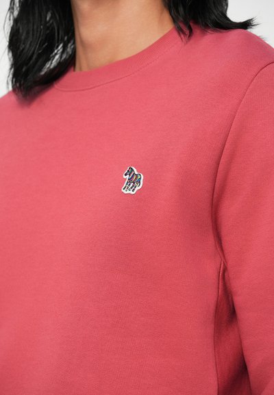 Rožinė sportinė palaidinė, pagaminta iš minkštos medžiagos, su apvaliu kaklu ir mažai, spalvinga auginio logotipu siuvinėtu ant krūtinės.