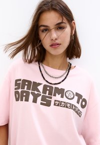 Lyserød bomulds T-shirt med korte ærmer, med "SAKAMOTO DAYS" i fed brunt skrift. Lagdelte perlekæder i varierede farver.