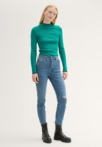 Langärmliges grünes Rippoberteil mit schwarzem Kragen, kombiniert mit hellblauen hochgeschnittenen Jeans mit einem gerissenen Knie und schwarzen Stiefeletten.