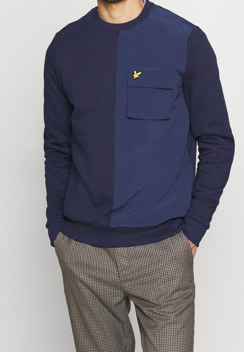 Homme portant une chemise à manches longues bleu marine et bleu foncé avec une poche poitrine et un pantalon marron à carreaux à motif.