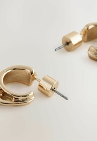 Goldene Metallohrringe mit glänzender Oberfläche. Kurvenform, zylindrische Stift-Details. Glatte Textur, mit einer Verschluss auf der Rückseite gesichert.