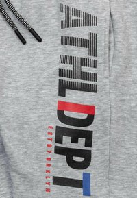 Pantalons de survêtement en mélange de coton gris avec du texte noir en gras, des accents rouges et bleus. Le design comprend des cordons décoratifs et une texture douce.