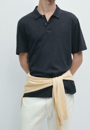 Personne portant un polo sombre à manches courtes avec un pull clair noué autour de la taille et un pantalon clair sur un fond uni.