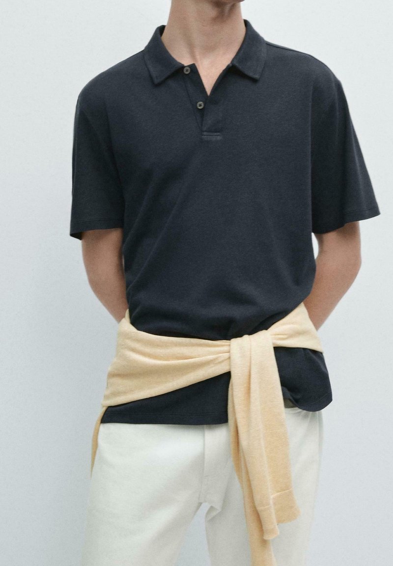 Personne portant un polo sombre à manches courtes avec un pull clair noué autour de la taille et un pantalon clair sur un fond uni.