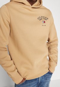 Beżowa bluza z kapturem wykonana z bawełny, z logo z przodu: "HILFIGER NEW YORK", z małym wzorem flagi. Posiada kieszeń kangurkę i ściągacze na mankietach.