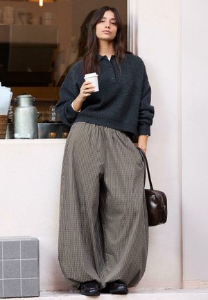 Nő a falnak dőlve, kávéscsészét tartva, sötét pulóvert, széles kockás nadrágot, fekete cipőt visel, és sötét kézitáskát tart a kezében.
