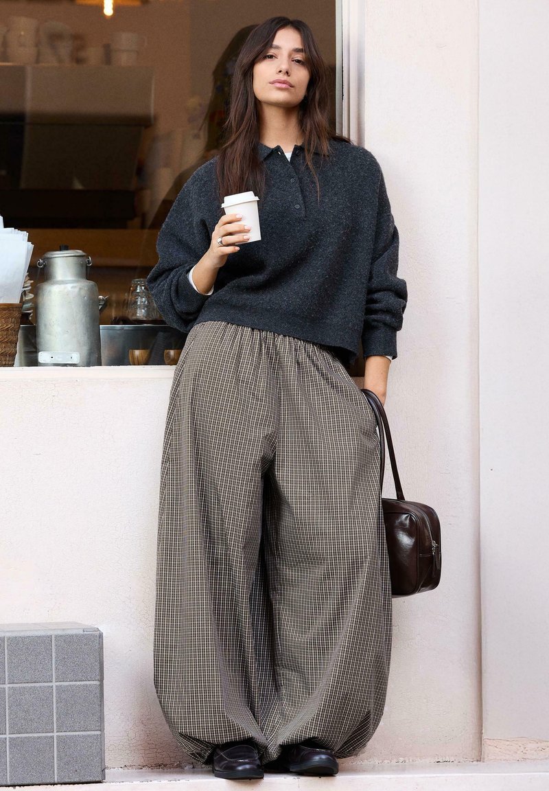 Femme appuyée contre un mur tenant une tasse de café, portant un pull sombre, un pantalon large à carreaux, des chaussures noires et tenant un sac à main foncé.